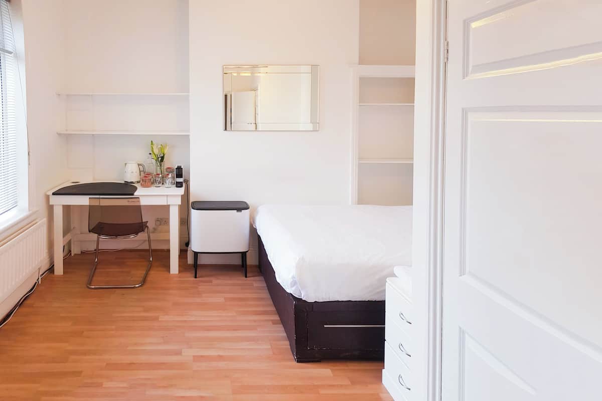 A bedroom with a bed, a desk, a chair, a small table with a lamp, and a mirror on the wall.