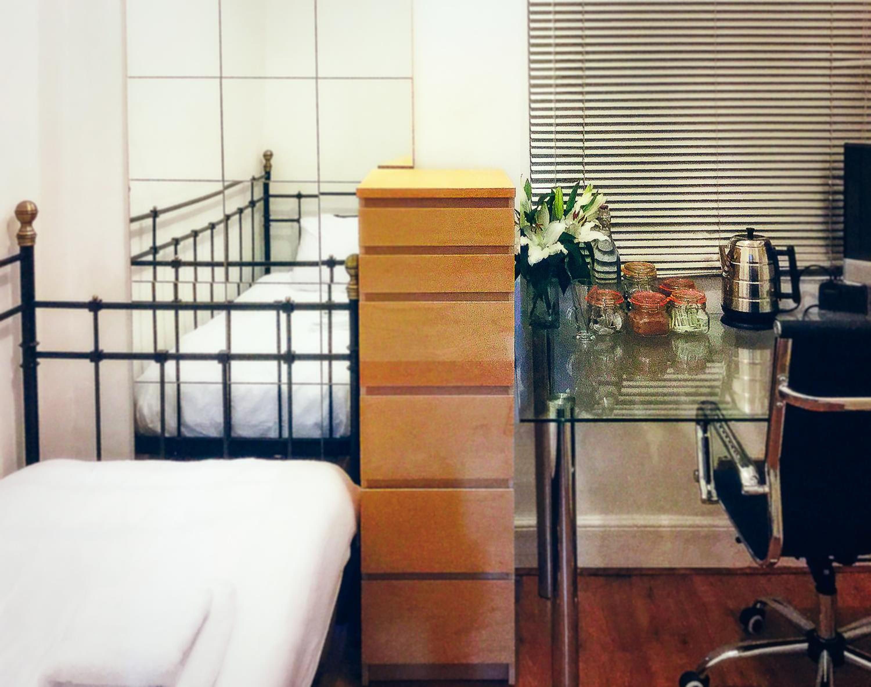Una habitación de hotel con una cama, una mesita de vidrio con un florero, una cafetera y una silla.