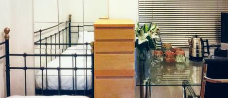 Una habitación de hotel con una cama, una mesita de vidrio con un florero, una cafetera y una silla.