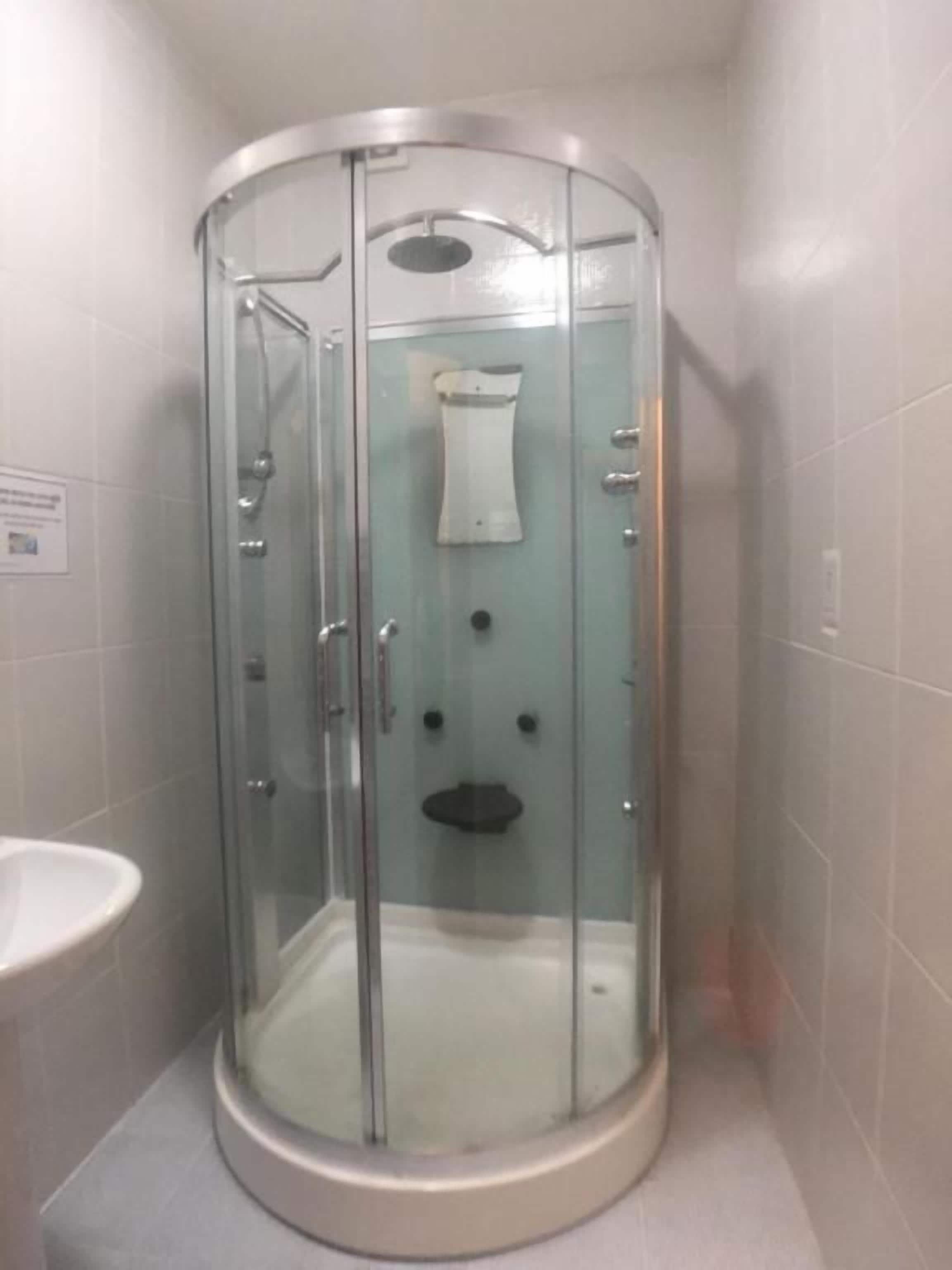 Un box de ducha con paredes de vidrio, una toalla blanca colgada en la puerta, un lavamanos a la izquierda y una pared con azulejos.