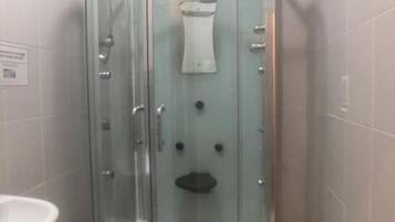 Un box de ducha con paredes de vidrio, una toalla blanca colgada en la puerta, un lavamanos a la izquierda y una pared con azulejos.