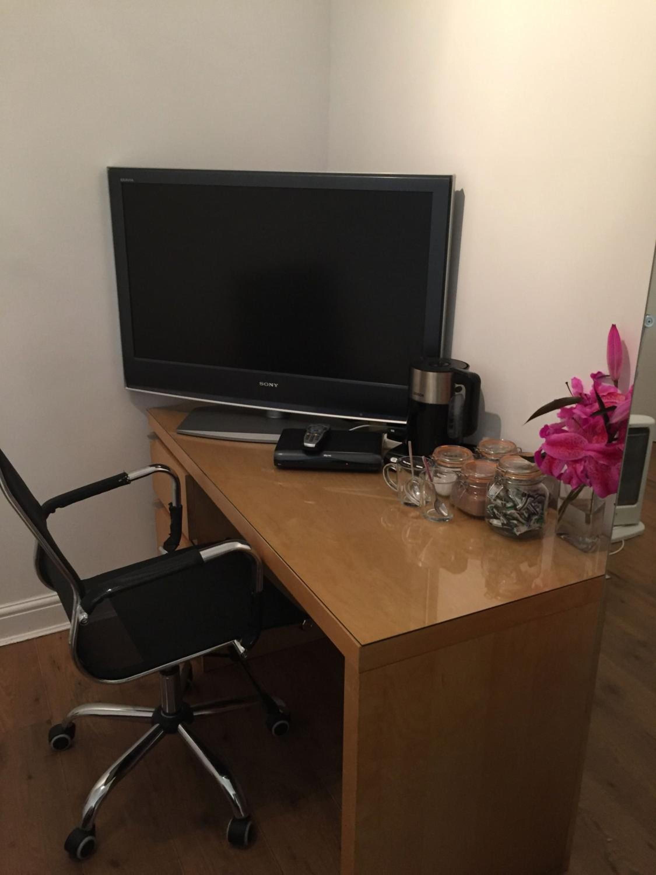 A workspace with a flat-screen TV, a black office chair, a wooden desk, a coffee maker, a vase with pink flowers, and jars with items on the desk.