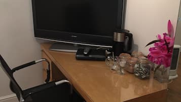 A workspace with a flat-screen TV, a black office chair, a wooden desk, a coffee maker, a vase with pink flowers, and jars with items on the desk.