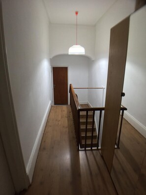 A narrow hallway with wooden flooring, a wooden staircase with a metal railing, and a white door at the end. - Outram 26 (Stockton-on-Tees)