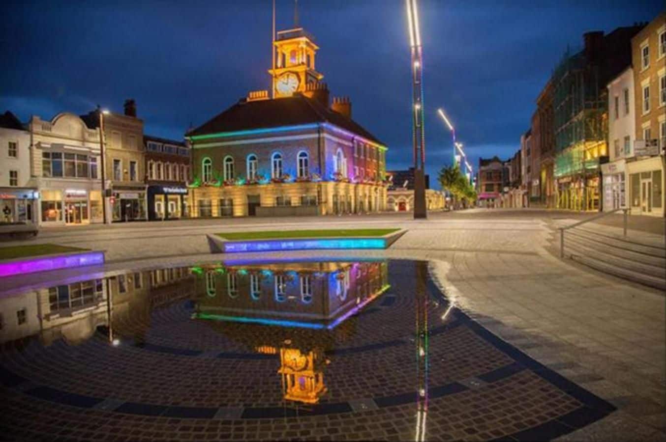 A square with a reflective water feature and a building with a clock tower.