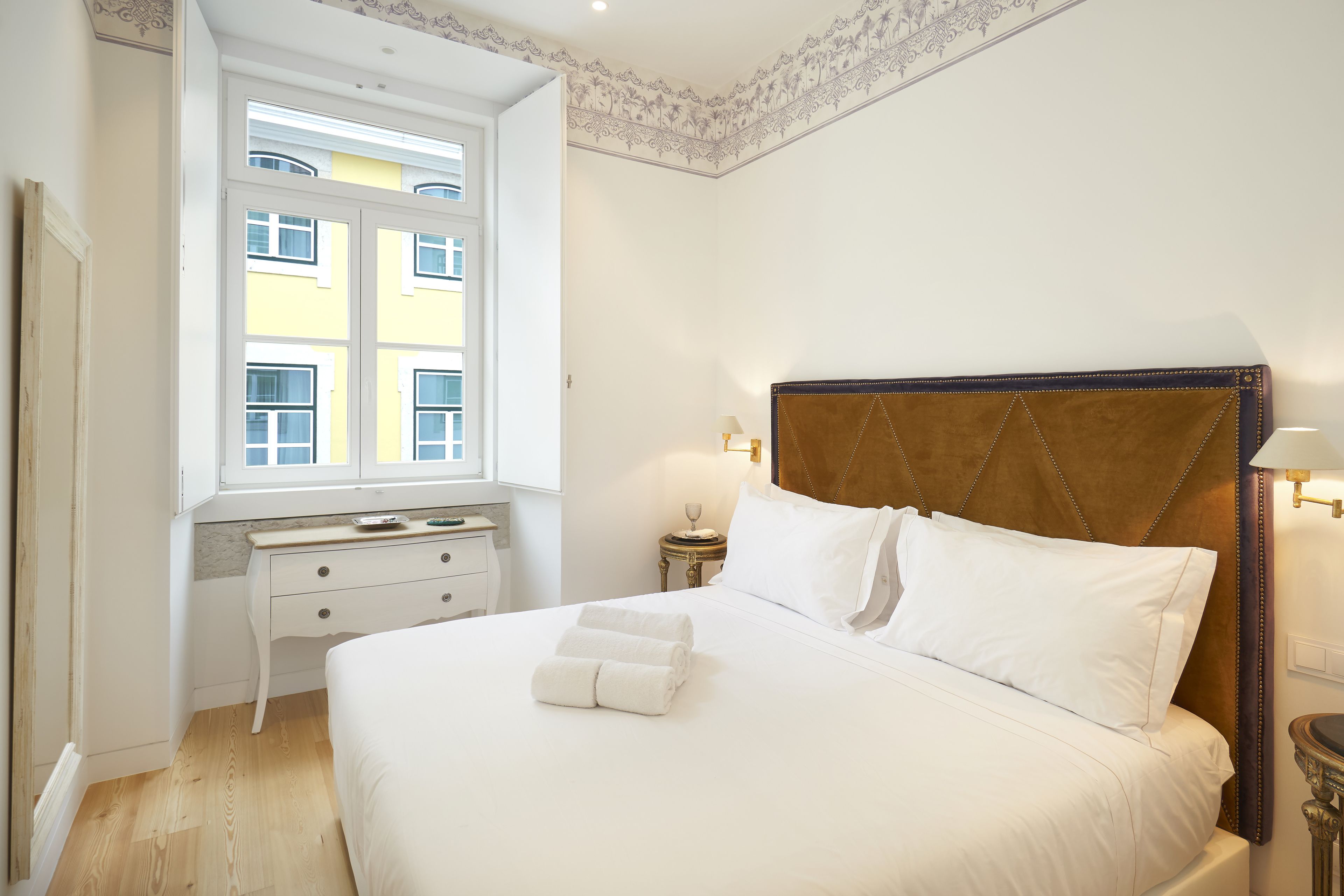 En pænt redt seng med hvide sengetæpper og to foldede håndklæder, et natbord med en lampe, og et vindue med udsigt til bygninger.