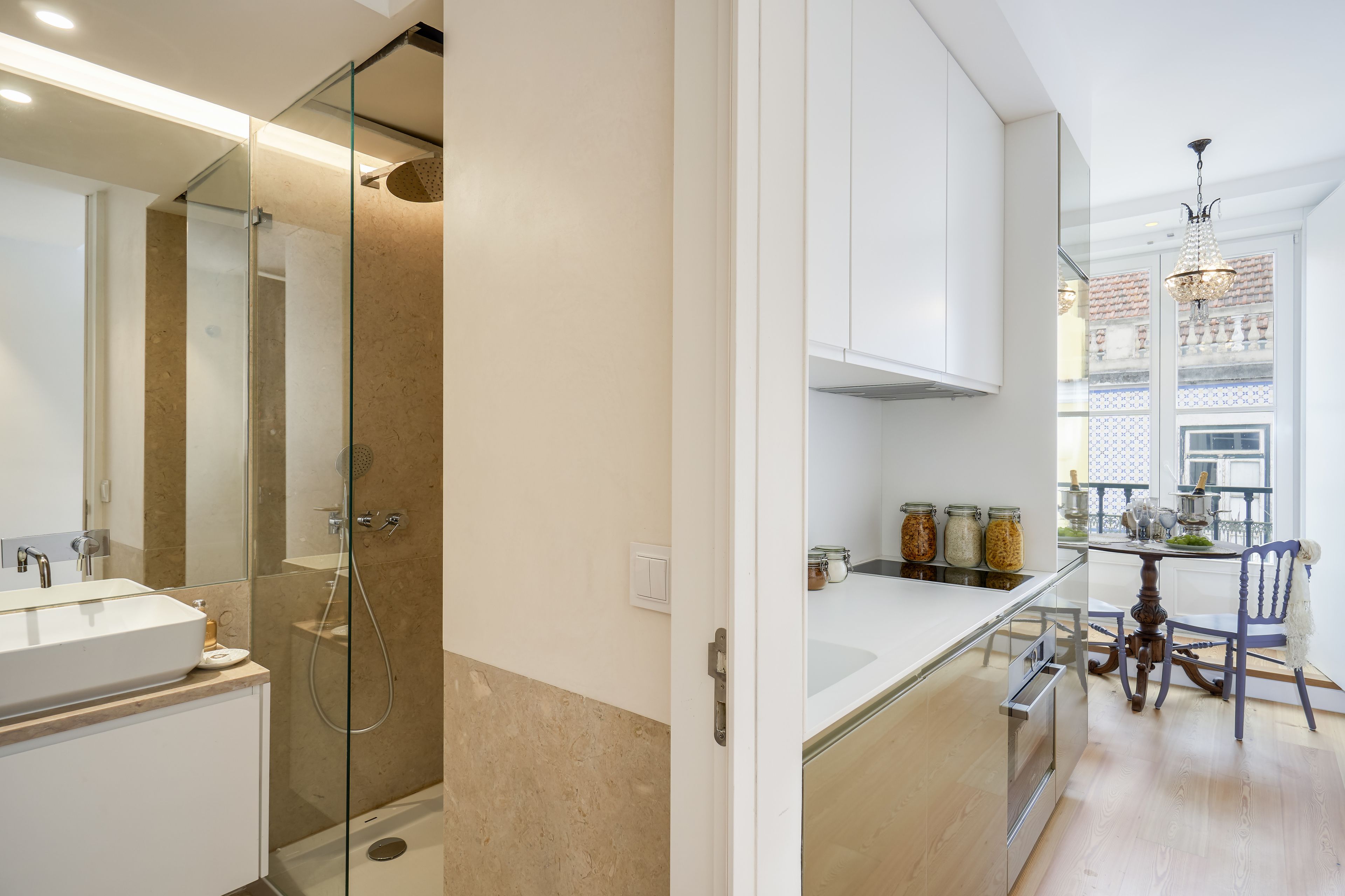 A modern bathroom with a glass shower enclosure, a white sink, and a mirror. Adjacent to the bathroom is a kitchen with a dining table, chairs, and a view of the city through a window.