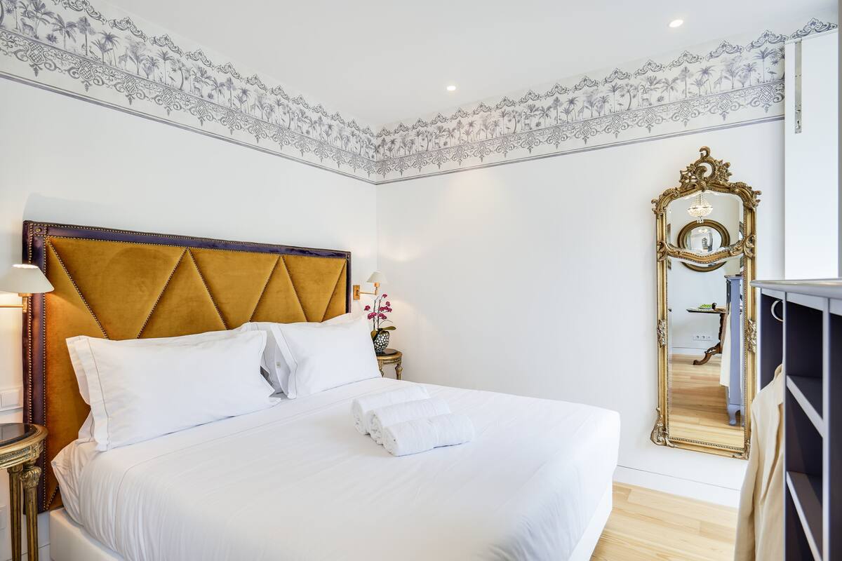 A neatly made bed with white linens, a gold-toned headboard, and a large ornate mirror.