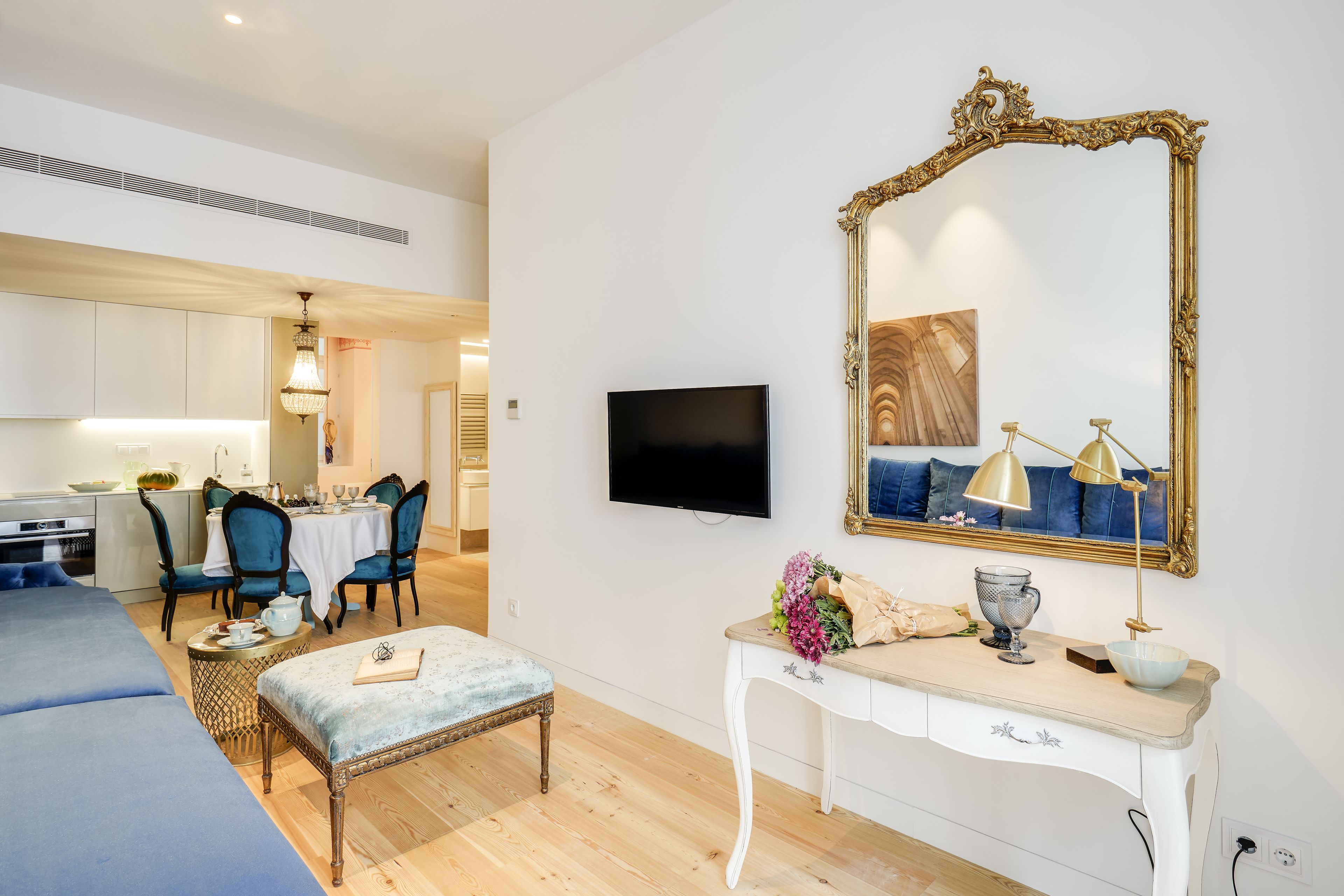 A modern living room with a flat-screen TV, a large ornate mirror, a dining area with blue chairs, and a kitchen in the background.