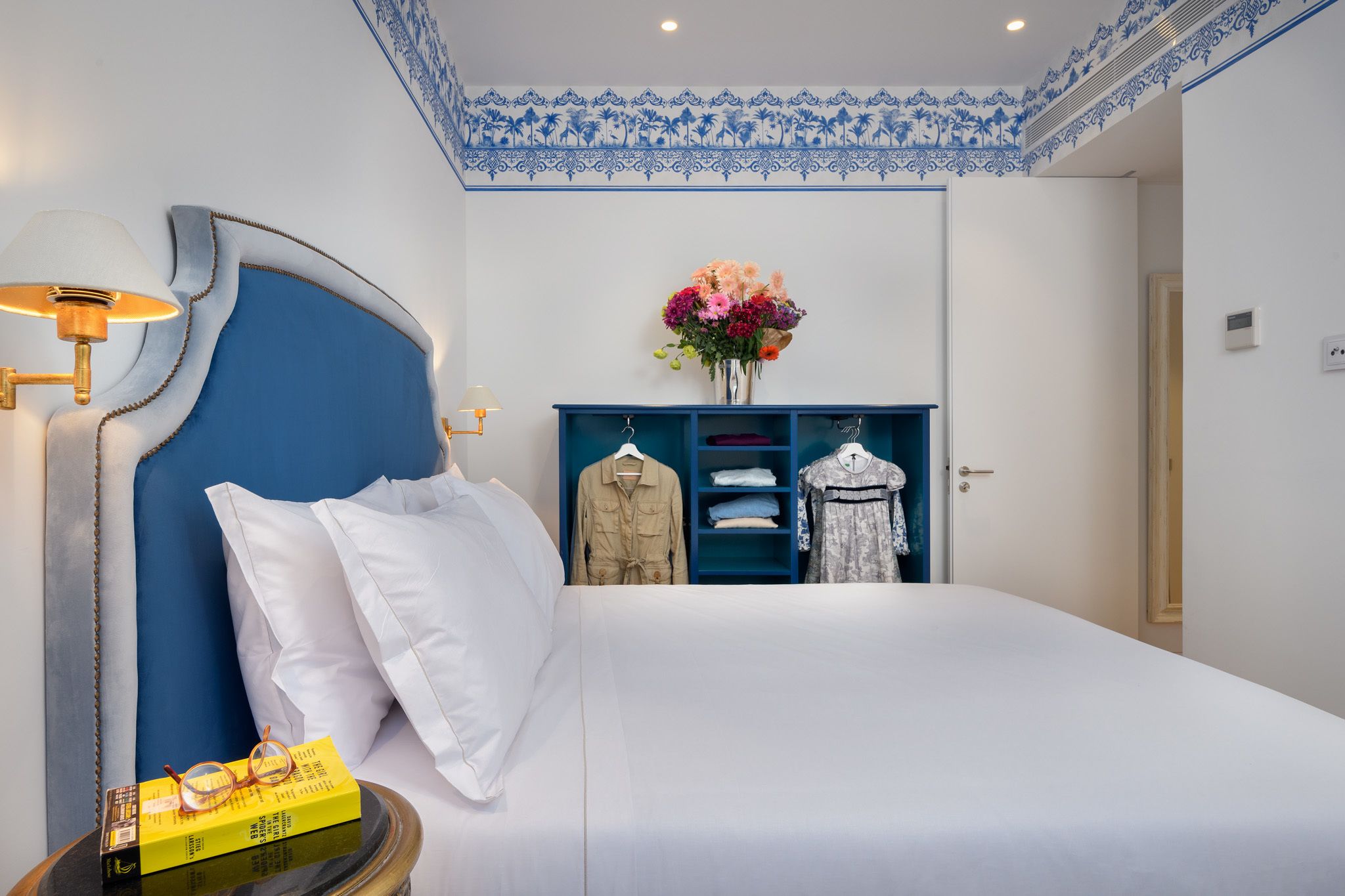 Une chambre d’hôtel bien rangée, avec un grand lit, une table de chevet sur laquelle sont posés des verres, une lampe et une armoire contenant des vêtements suspendus.