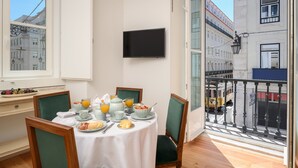 A dining area with a table set for breakfast, a TV mounted on the wall, and a view of the city through large windows. - Residentas Aurea (Lisbon)