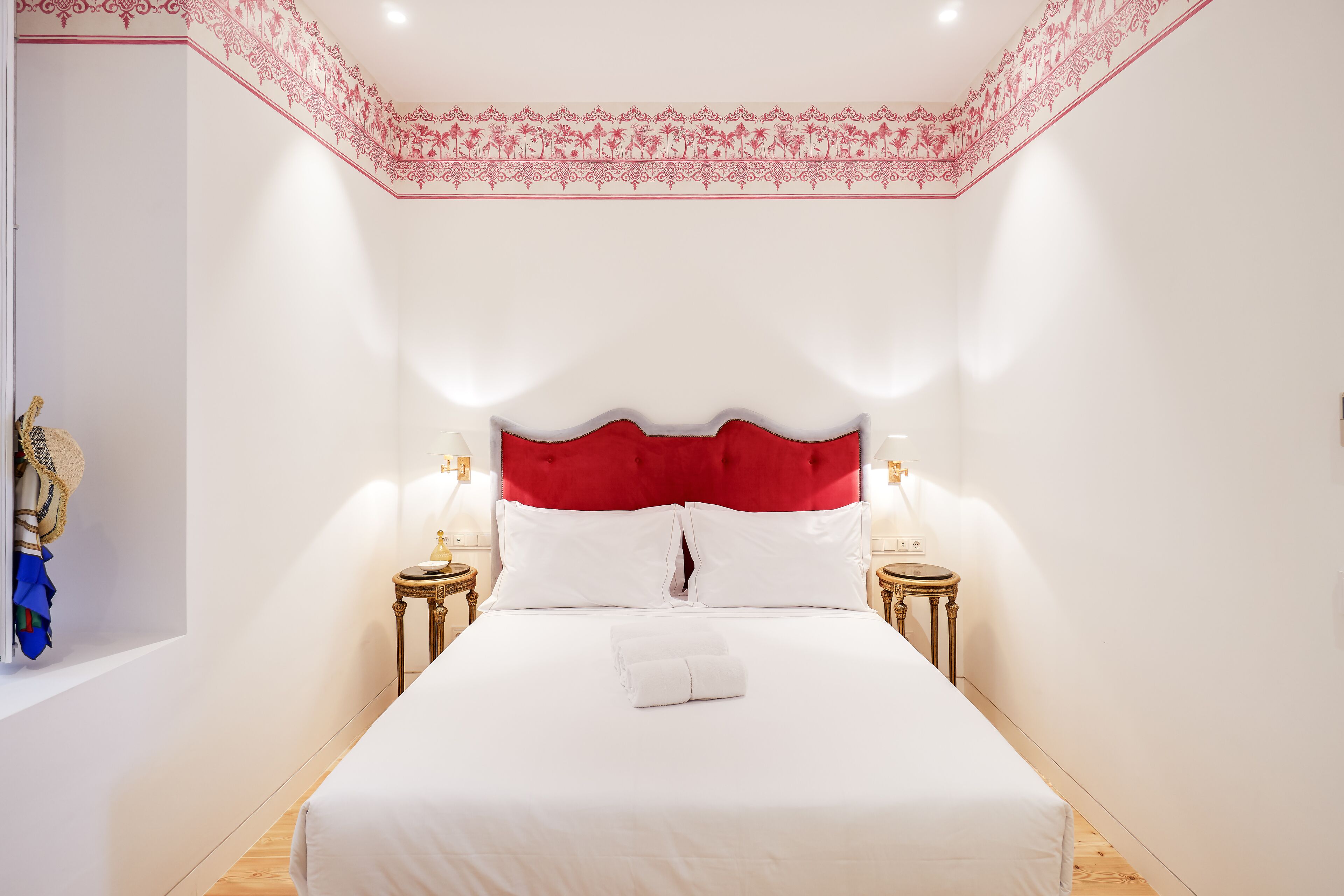 A hotel room with a large bed, white bedding, a red upholstered headboard, two bedside tables with lamps, and a decorative ceiling border.