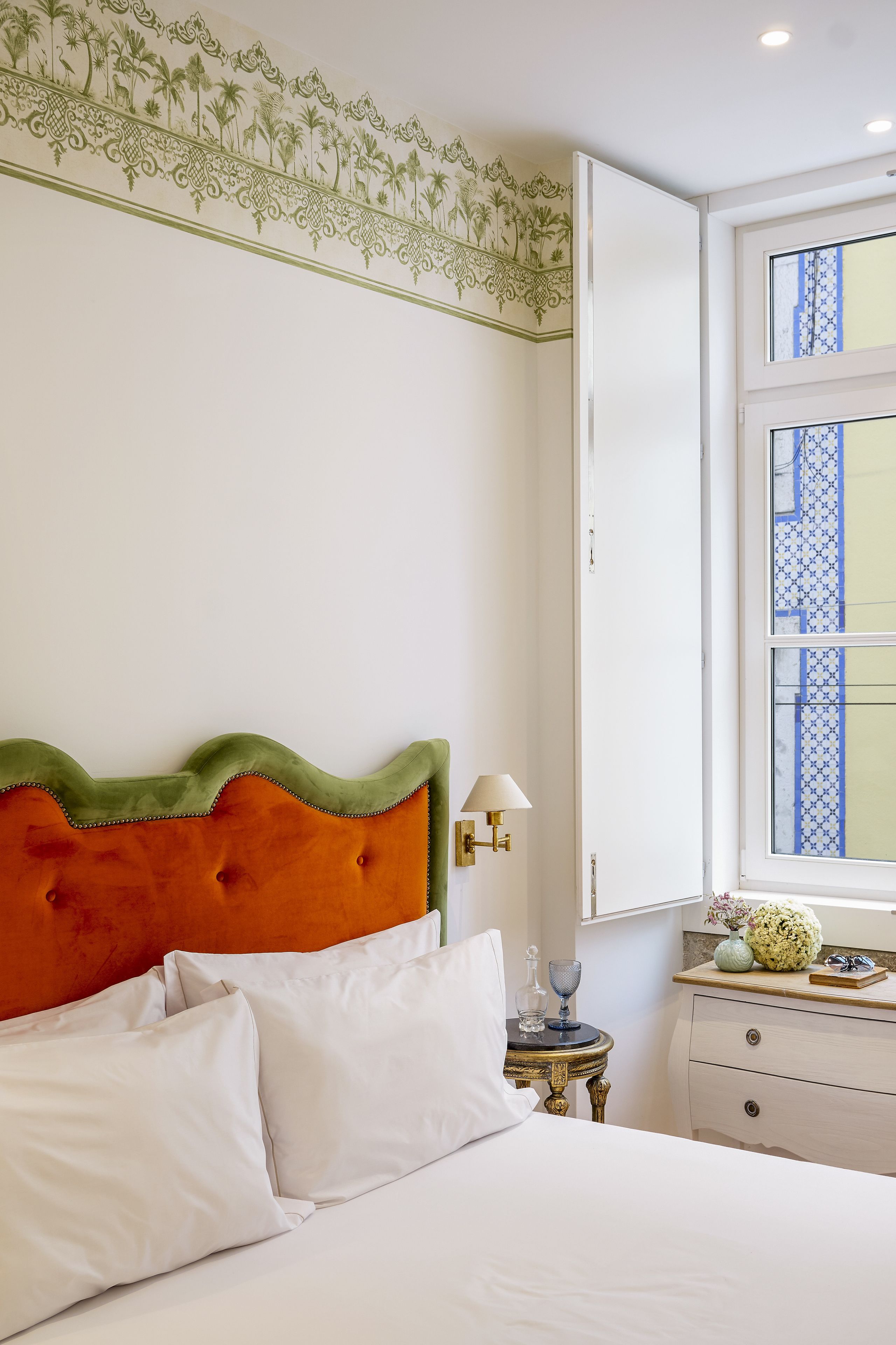 A neatly made bed with white and orange bedding, a small bedside table with a lamp, and a window with blue patterned curtains.