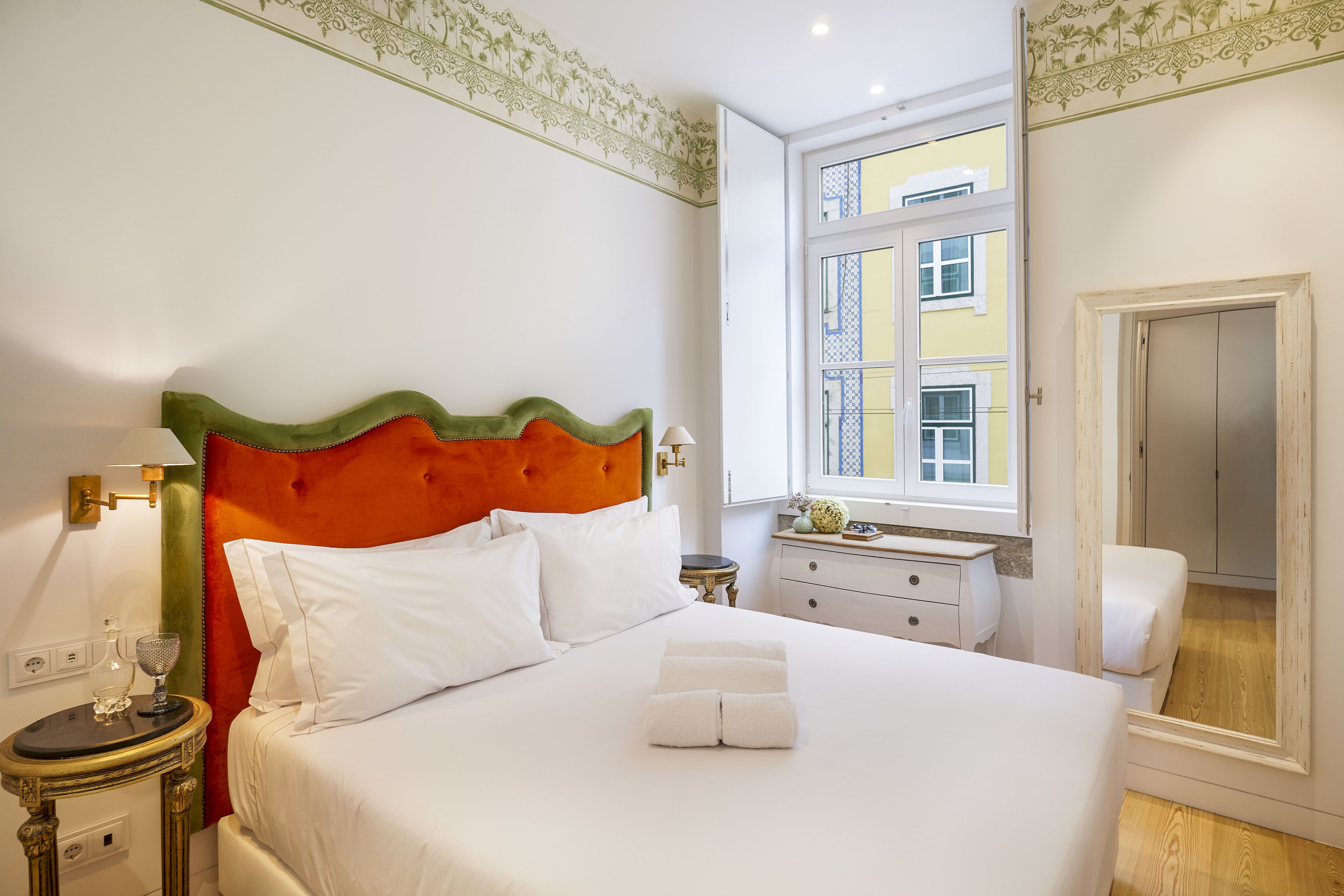 A neatly made bed with white linens and a folded towel, a bedside table with a lamp, and a window with a view of buildings.