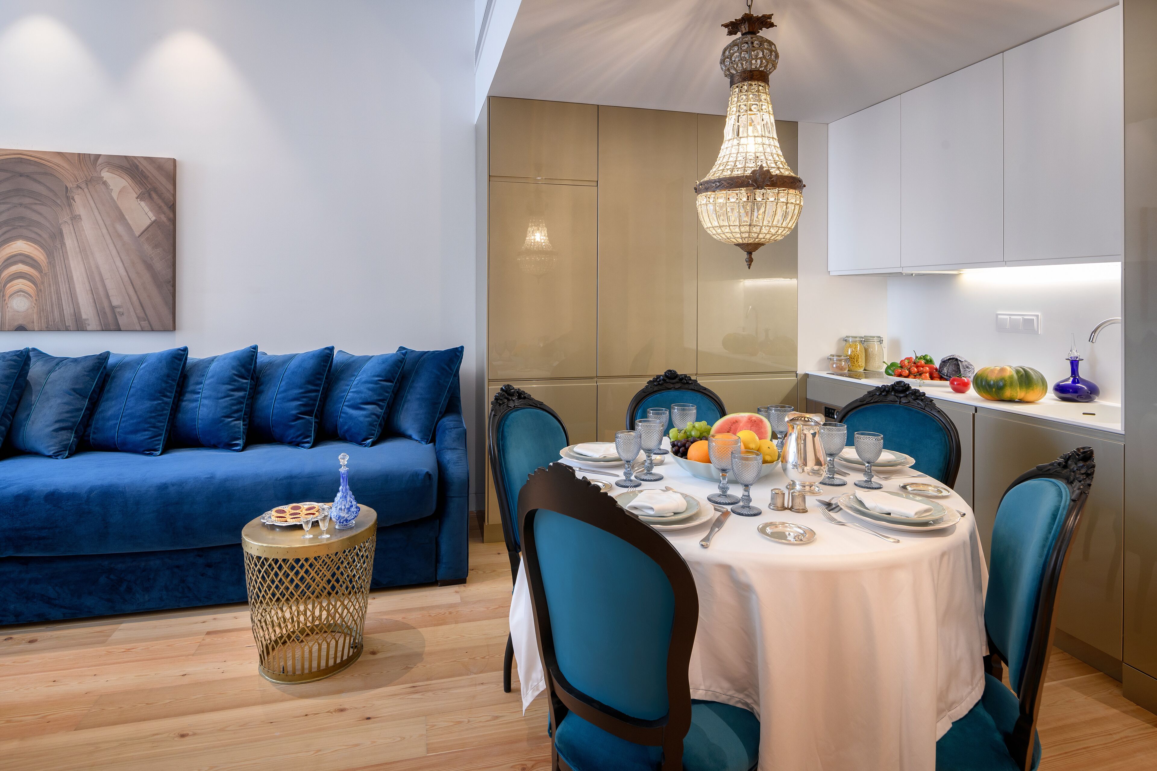 A modern dining area with a round table set for six, blue upholstered chairs, a blue sofa, and a chandelier.