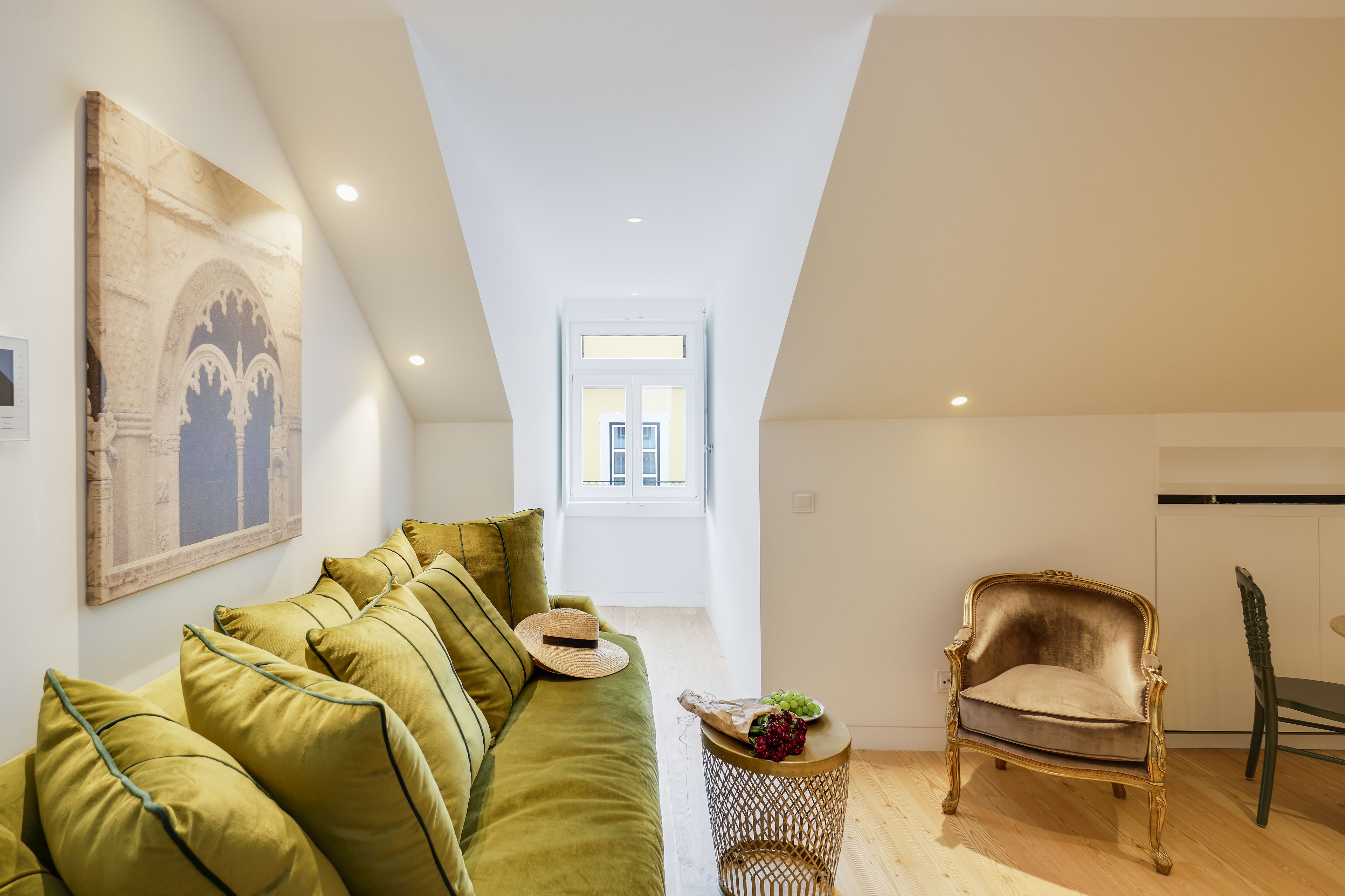 Un dormitorio tipo loft moderno con techo alto, un sofá de terciopelo verde, un sillón antiguo y un ventanal que proporciona luz natural.