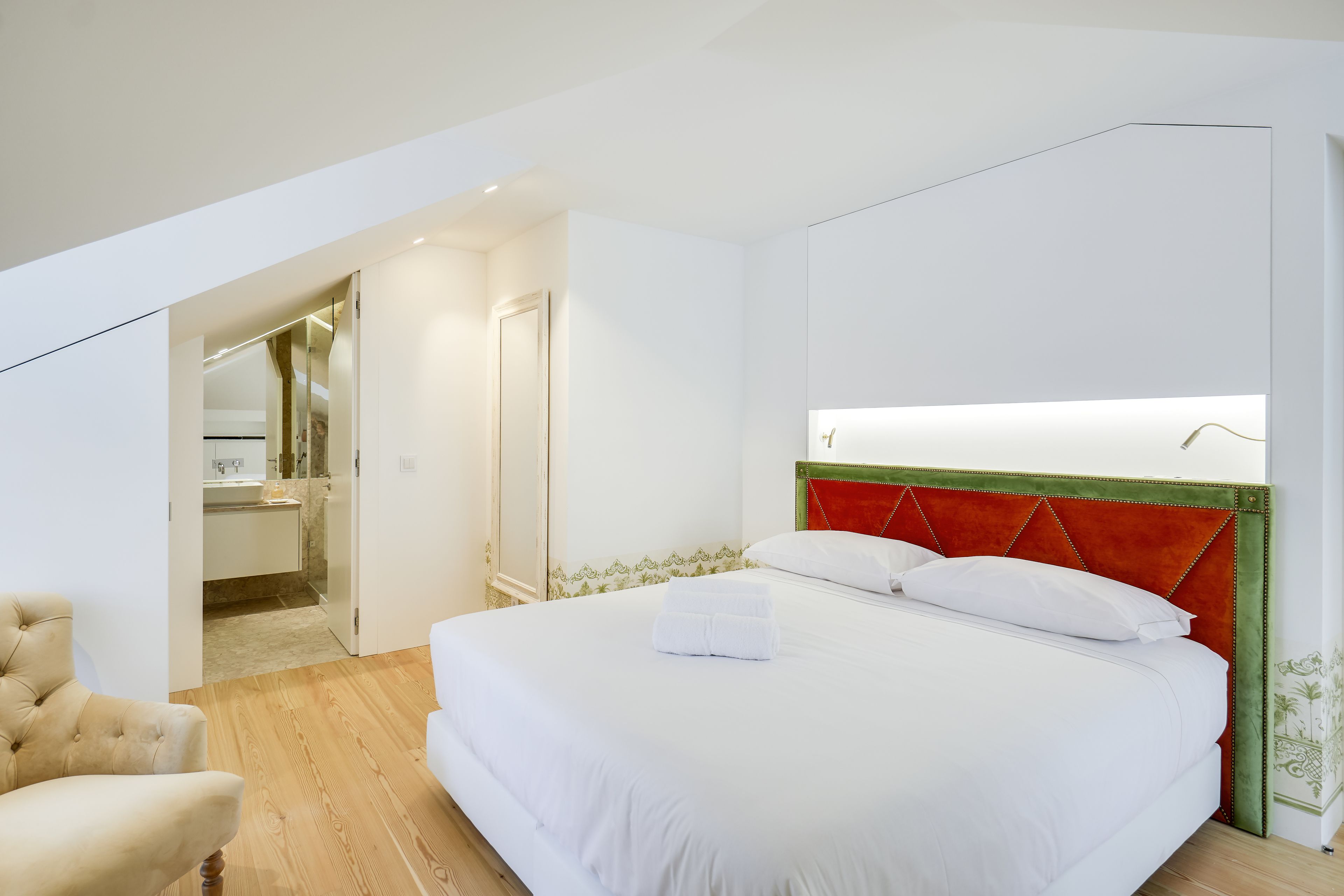 Een moderne hotelkamer met een groot bed, een houten vloer, een witte fauteuil en een badkamer zichtbaar door een open deur.