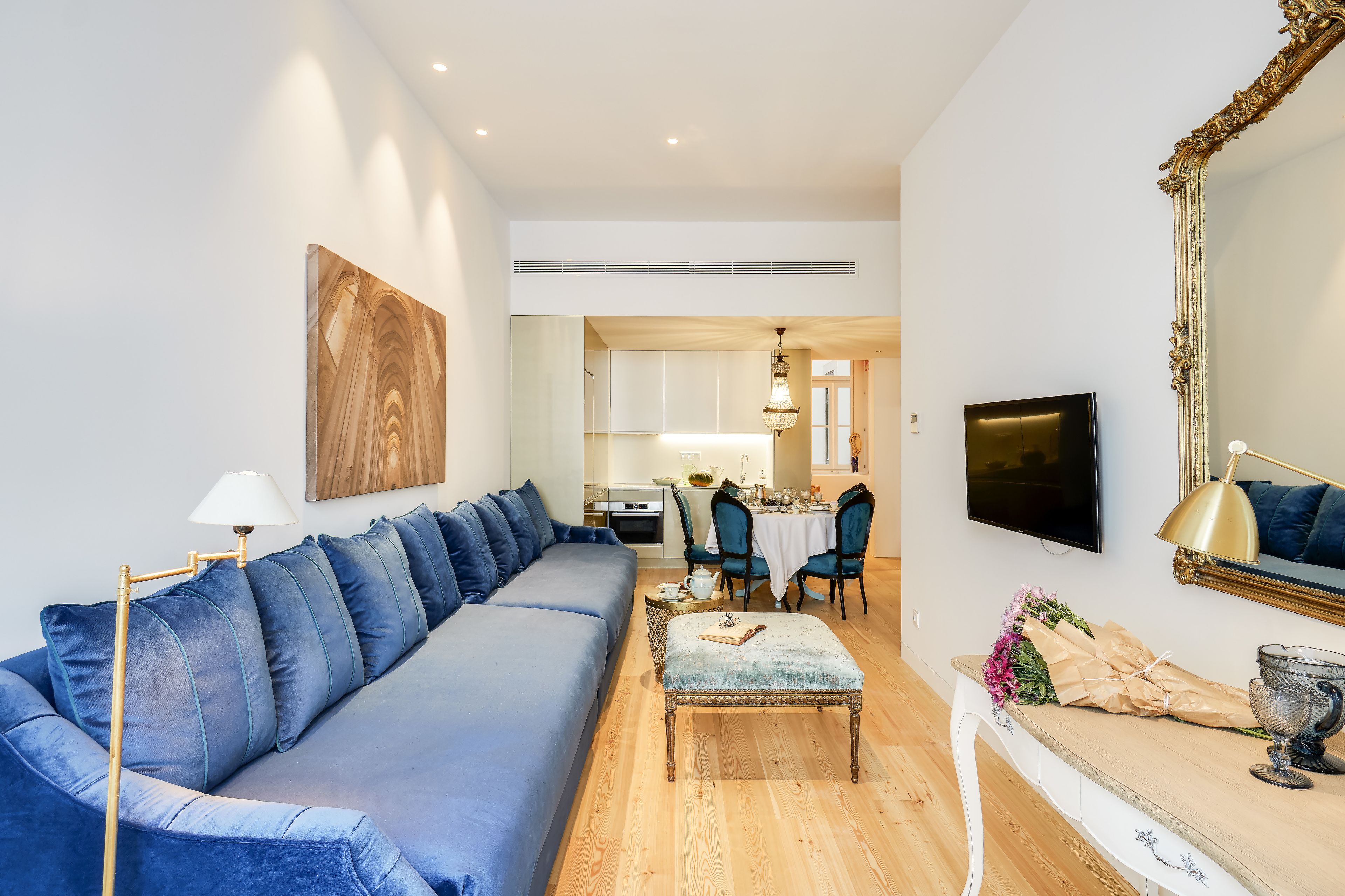 A modern living room with a blue sofa, a dining area with chairs, a TV, and a large mirror.