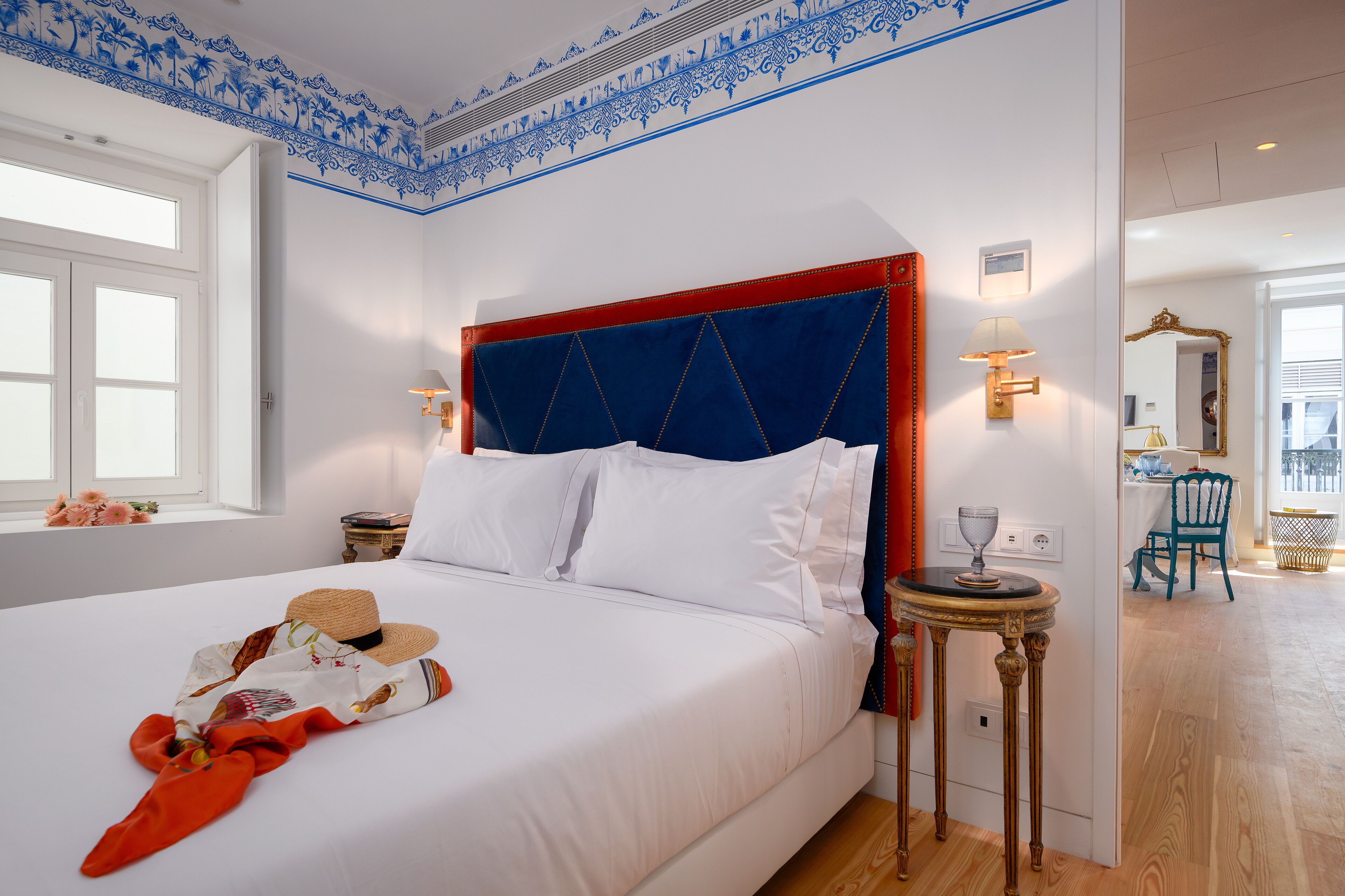 Una cama bien hecha con sábanas blancas y un cabecero rojo, una mesita redonda con una lámpara, y una ventana con vistas al exterior.