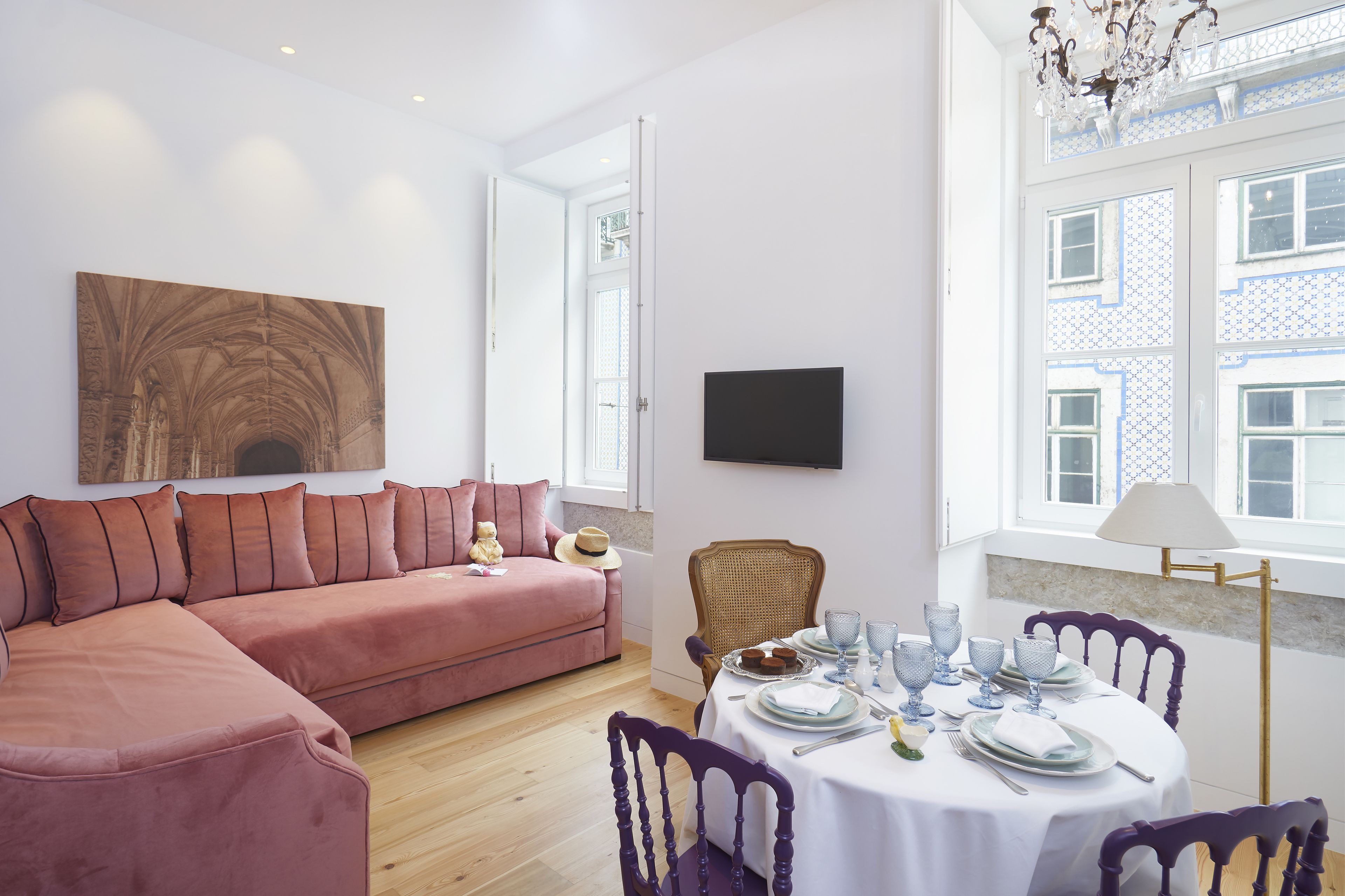 A modern living room with a pink sectional sofa, a dining table set with dinnerware, and a large window with patterned shutters.