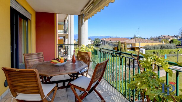 A balcony with a wooden table and chairs, a bowl of fruit, and a view of the surrounding area. - Desenzanoloft – Butterfly and Pools (Desenzano del Garda)