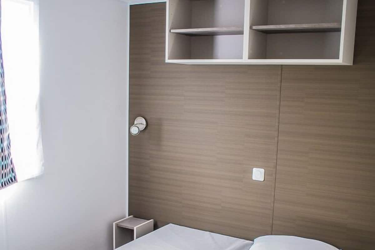 A neatly made bed with a green blanket and white pillows, a bedside table, and built-in shelves above the bed.
