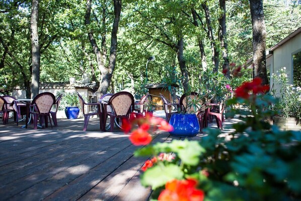 An outdoor patio area with wooden flooring, multiple tables and chairs, and potted plants.