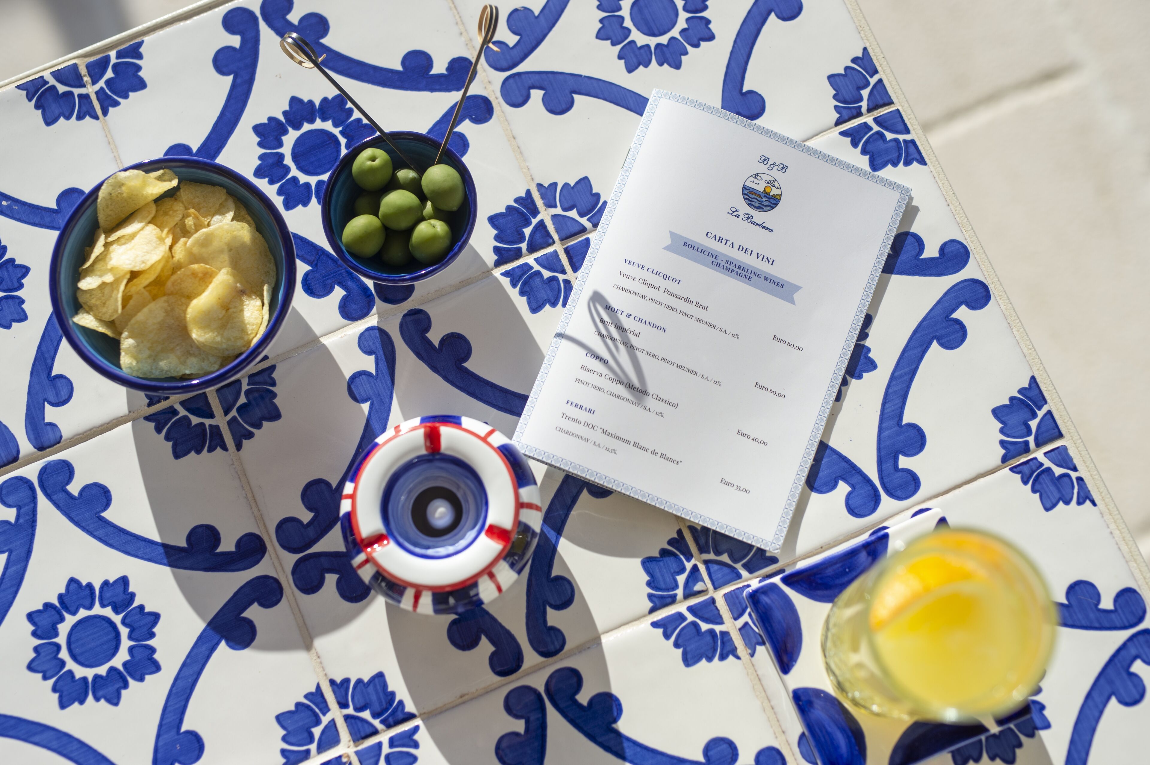 Una mesa con un estampado de baldosas azules y blancas, un cuenco de patatas fritas, un cuenco de aceitunas, un menú y una copa de vino.