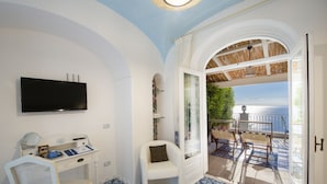 A room with a TV mounted on the wall, a desk with a chair, a small white chair, and a view of the sea through an open door. - B&B La Barbera (Praiano)