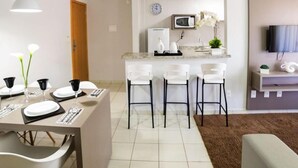 A modern kitchen with a microwave, a clock, and a small plant. There is a dining area with a table set for four and a bar with three stools. - Serra Madre Residence (Rio Quente)