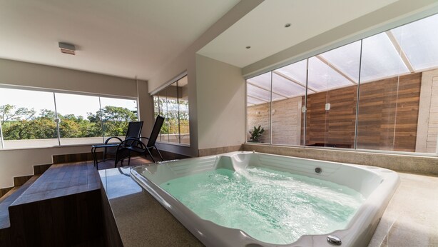 A modern indoor swimming pool with a wooden deck, a lounge area, and large windows offering a view of the outdoors. - Serra Madre Residence (Rio Quente)
