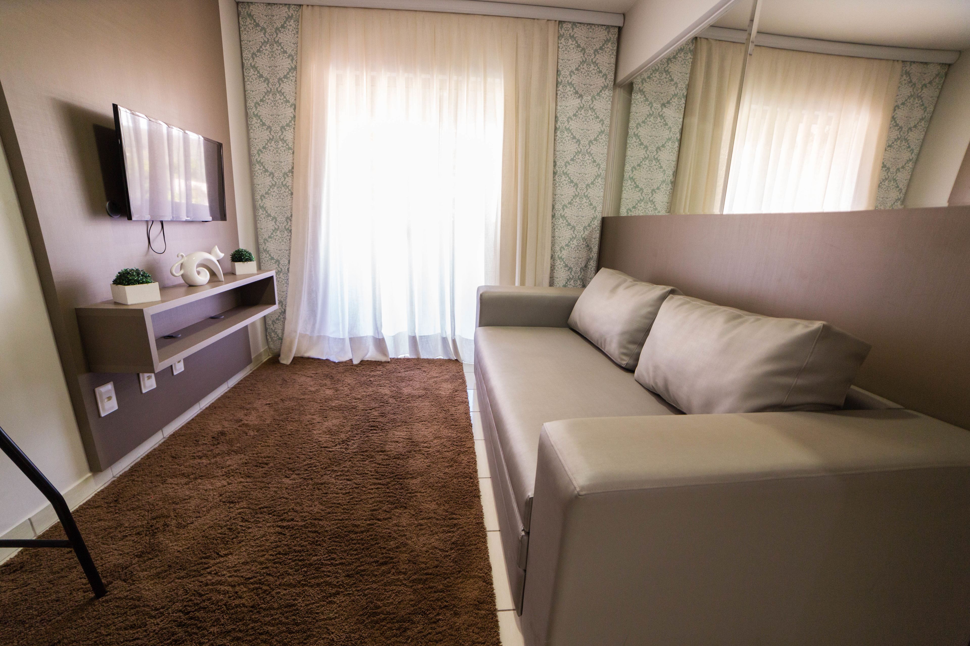 A modern living room with a beige sofa, a mounted TV, a small shelf with decorative items, and a patterned curtain.