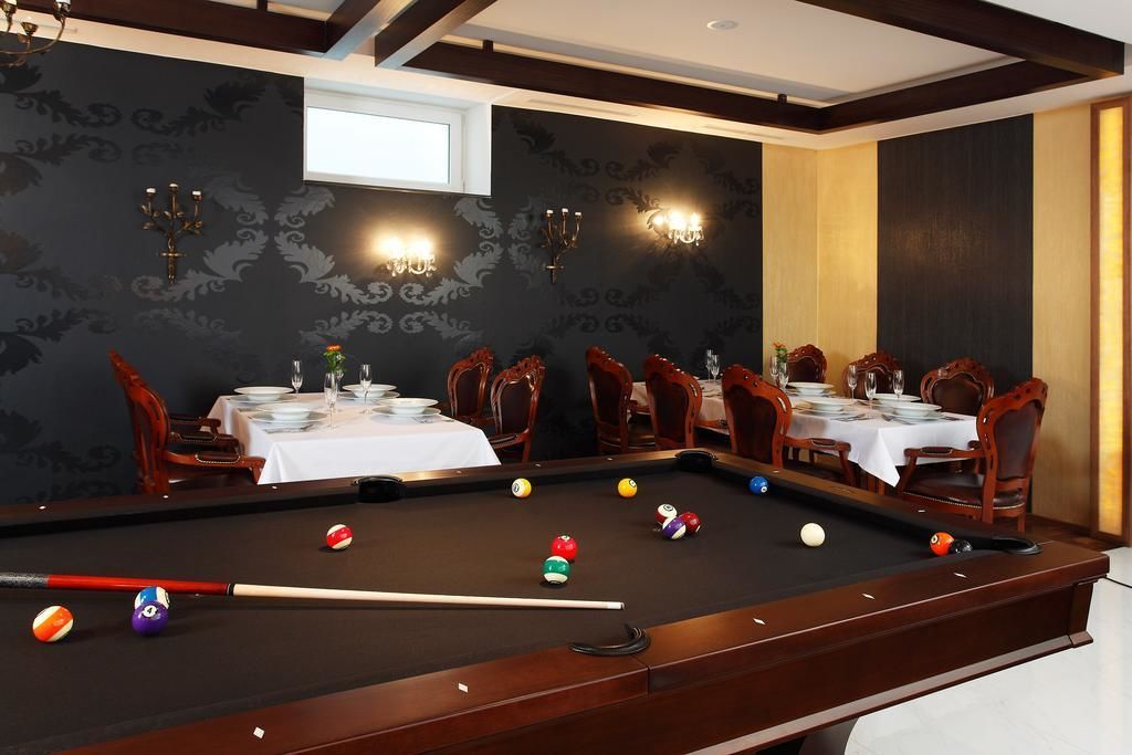 A pool table with balls, surrounded by dining tables and chairs, in a room with patterned wallpaper and wall sconces.