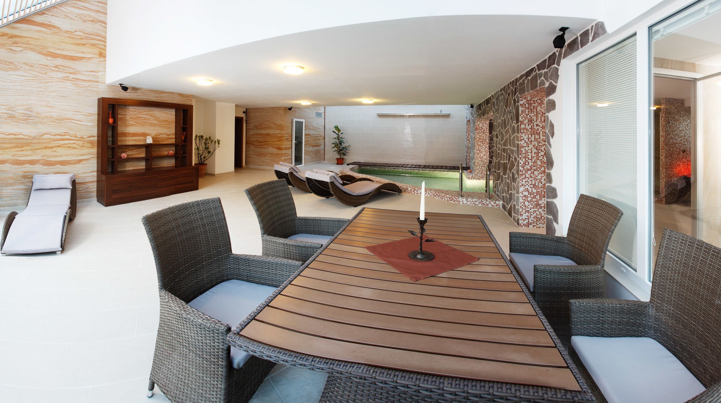 A modern outdoor lounge area with wicker furniture, a wooden table, and a stone wall with mosaic tiles.