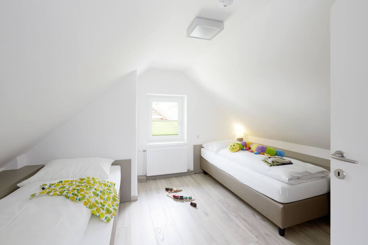 A small, well-lit bedroom with a sloped ceiling, a window, a bed with a white mattress and a colorful blanket, and a bedside table with toys.