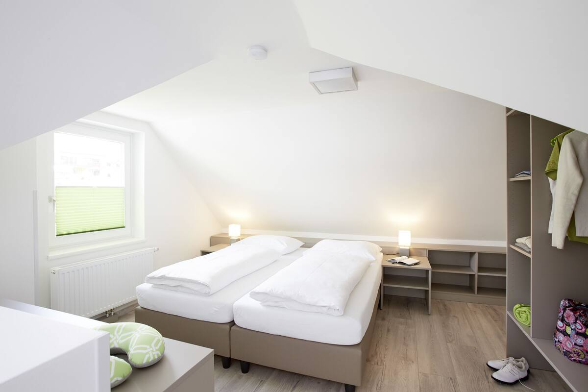 A bedroom with two beds, a window, a radiator, a bedside table with a lamp, a wardrobe, and a shoe rack.