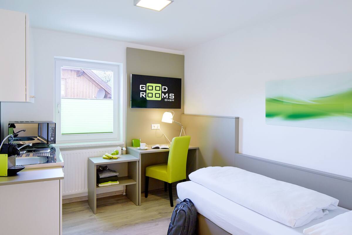 A hotel room with a bed, a desk with a computer, a chair, a radiator, and a TV on the wall.