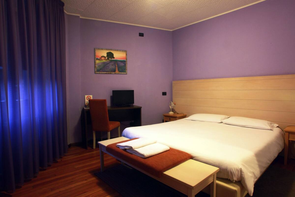 A hotel room with a large bed, a desk with a computer, a chair, and a painting on the wall.