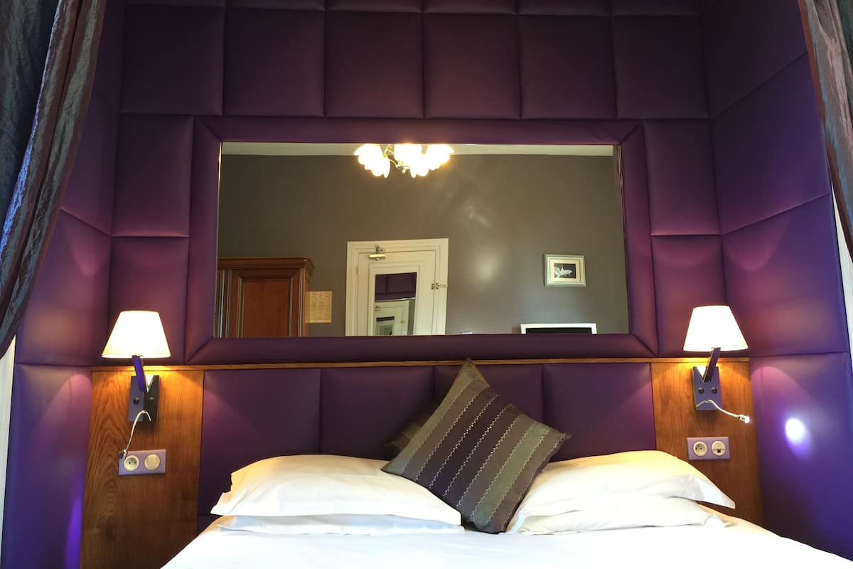A bedroom with a large mirror, two bedside lamps, a bed with white linens, and a purple headboard.