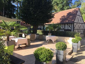 Un espace extérieur aménagé en terrasse, avec des tables et des chaises, entouré de plantes en pot et d’un grand arbre.