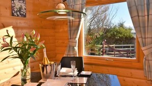 A wooden cabin interior with a dining table set for two, a vase with flowers, and a bottle of champagne. - Pheasant Lodge Scottish Borders (Carlisle)
