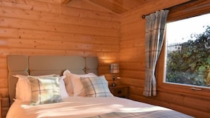 A wooden cabin bedroom with a bed, pillows, a nightstand, and a window with curtains. - Pheasant Lodge Scottish Borders (Carlisle)