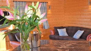 A cozy cabin interior with a wooden log wall, a dining table with a vase of flowers, a dark brown sofa with cushions, and a window with curtains. - Pheasant Lodge Scottish Borders (Carlisle)
