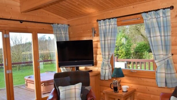 A cozy cabin interior with a wooden interior, a flat-screen TV mounted on the wall, and a comfortable seating area with a table and a lamp. - Pheasant Lodge Scottish Borders (Carlisle)