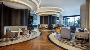 Un hall d’entrée spacieux doté d’un plancher en bois incurvé, d’un espace de détente central avec des canapés et des fauteuils, et de grandes fenêtres offrant une vue sur l’extérieur.