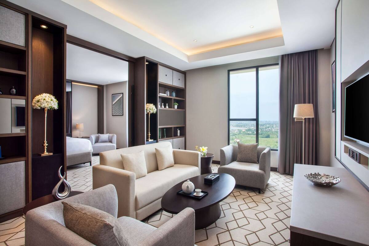 Een moderne hotelkamer met een bank, fauteuils, een salontafel en een televisie. Er is een groot raam met uitzicht op de stad.