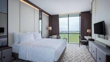 A hotel room with a large bed, a TV, a desk with a chair, and a view of the city through large windows.