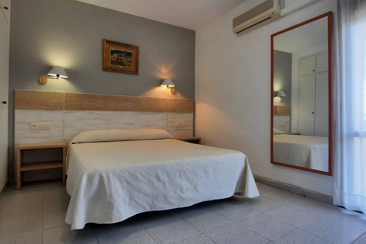 Een hotelkamer met een bed, nachtkastje, een wandlamp, een lijst met afbeelding en een grote spiegel.