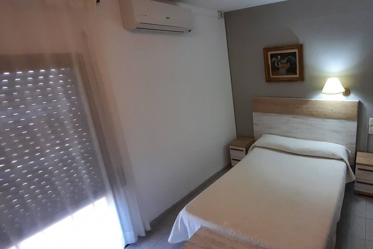 Een hotelkamer met een bed, nachtkastjes, een lamp, een airconditioning en een schilderij aan de muur, plus een raam met zonwering.