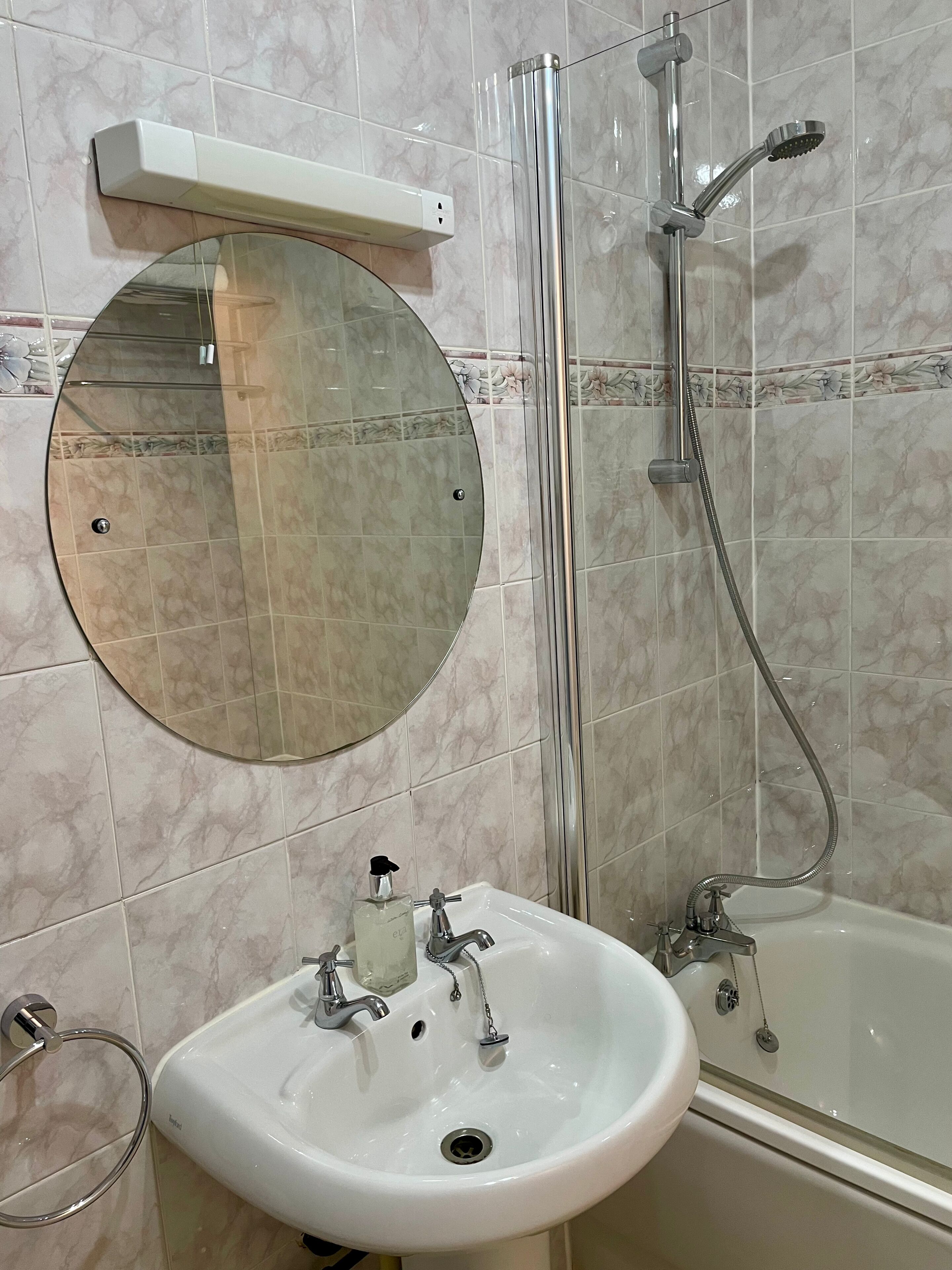 A bathroom with a round mirror, a sink with soap dispenser, and a shower with a handheld showerhead.